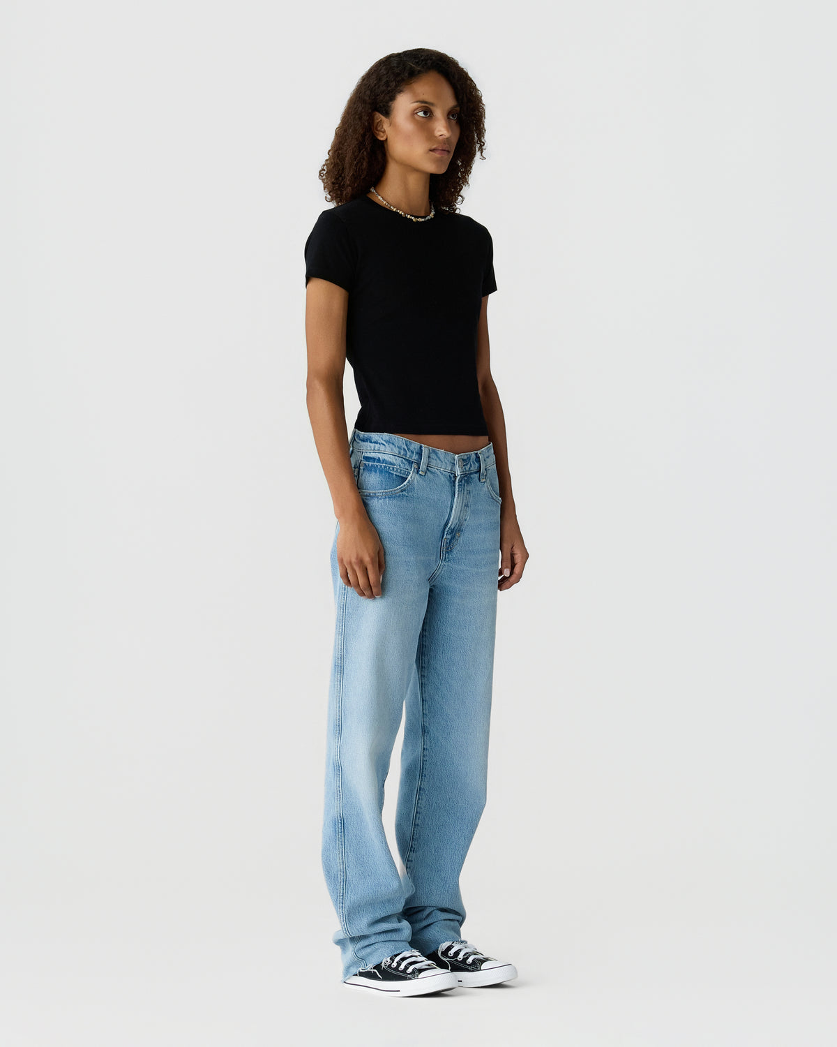 Woman with curly hair wearing a black short sleeve top, light blue wide leg jeans, and black and white sneakers against a light gray background