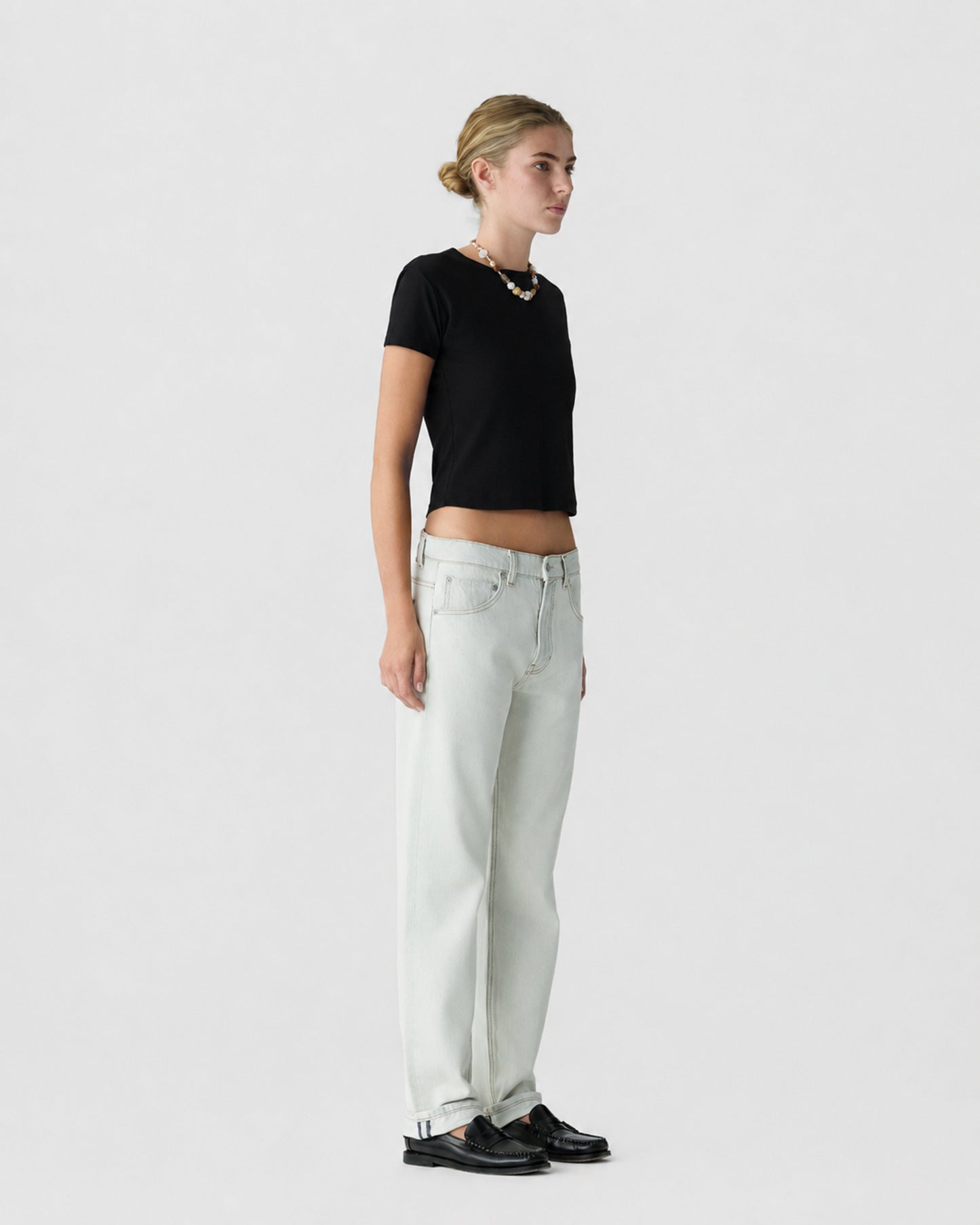 Woman standing in profile wearing a black short sleeve top, light gray wide leg jeans, black loafers, and a necklace against a light gray background