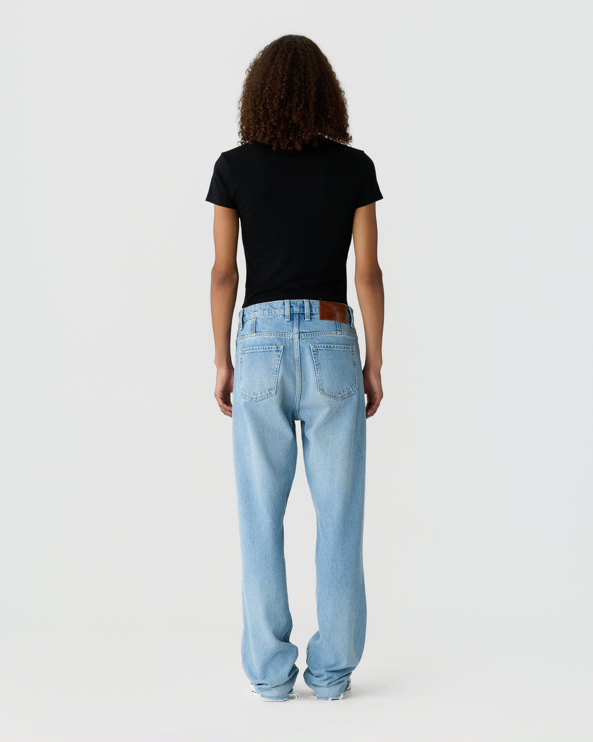 Back view of a person wearing a black short sleeve T shirt, light blue straight leg jeans, and gray sneakers against a plain light background