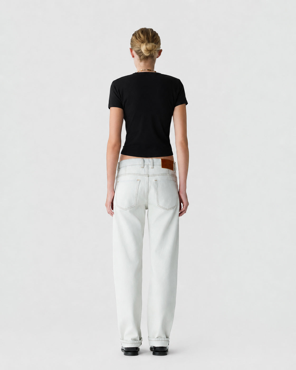 Rear view of a person wearing a black short sleeve shirt, white high waisted jeans, and black shoes against a light gray background