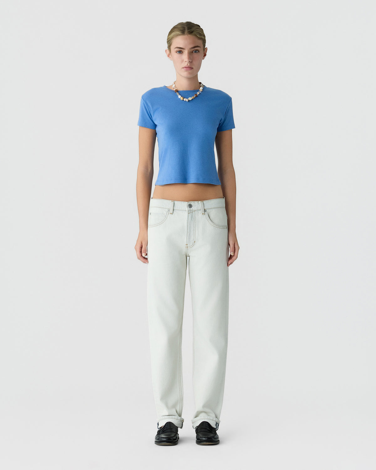 Model wearing a blue cropped top, white high-waisted jeans, black shoes, and a beaded necklace against a light gray studio background.
