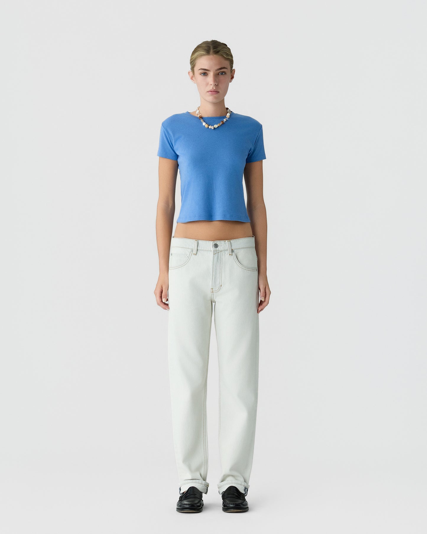 Model wearing a bright blue cropped short sleeve top, white wide leg jeans, black shoes, and a beaded necklace against a light gray background