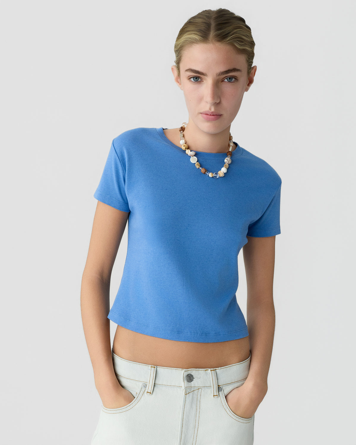 Woman wearing a bright blue short sleeve top with white jeans and a beaded necklace against a light gray background