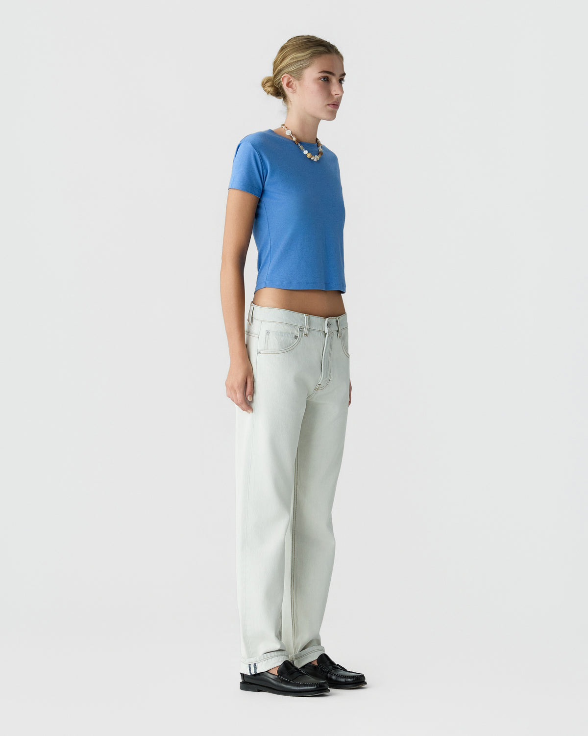 Model standing in profile wearing a bright blue short sleeve top, light wash wide leg jeans, layered necklaces, and black loafers against a white background