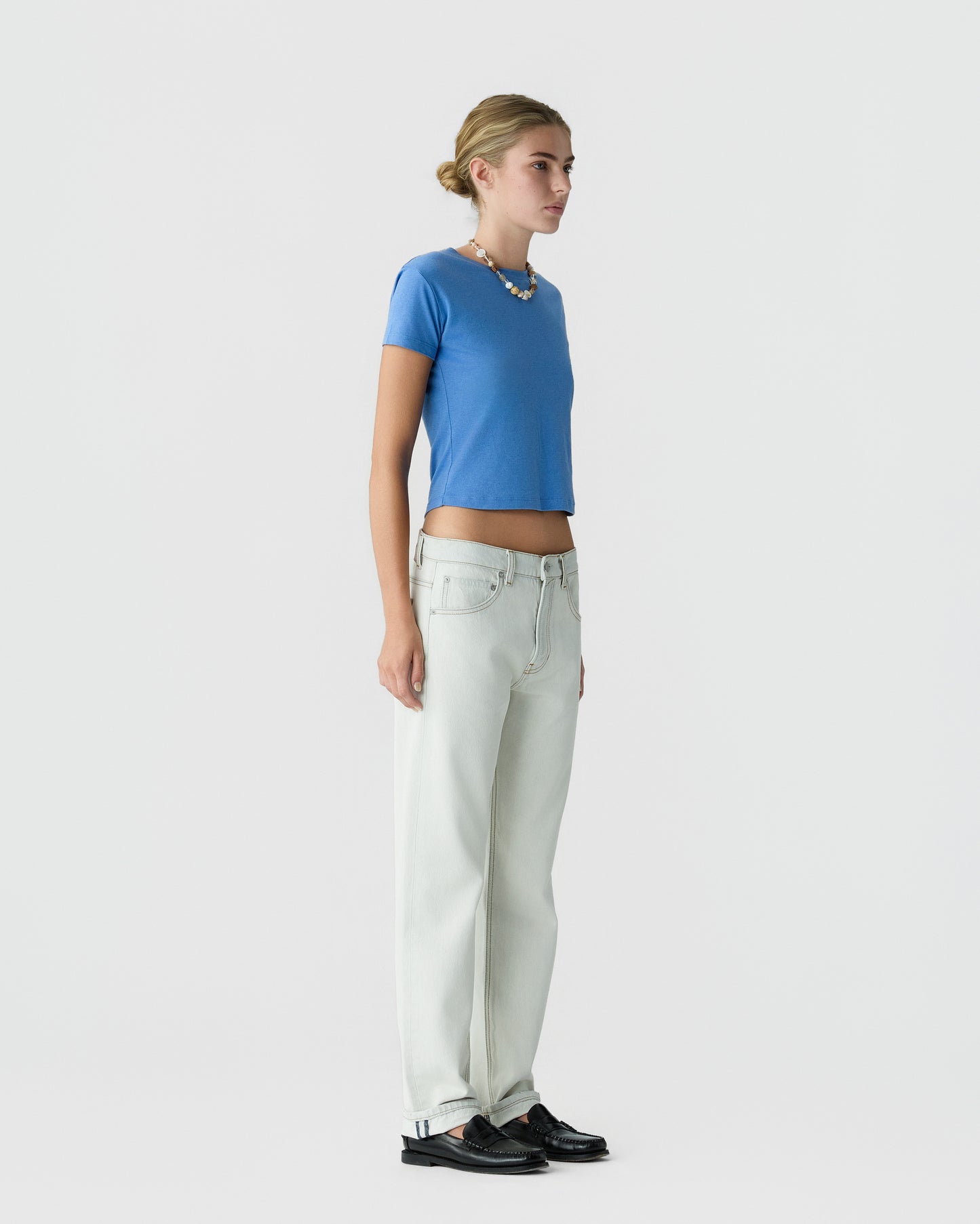 Woman standing in profile wearing a blue short sleeve top with pale wide leg trousers and black loafers against a light gray background