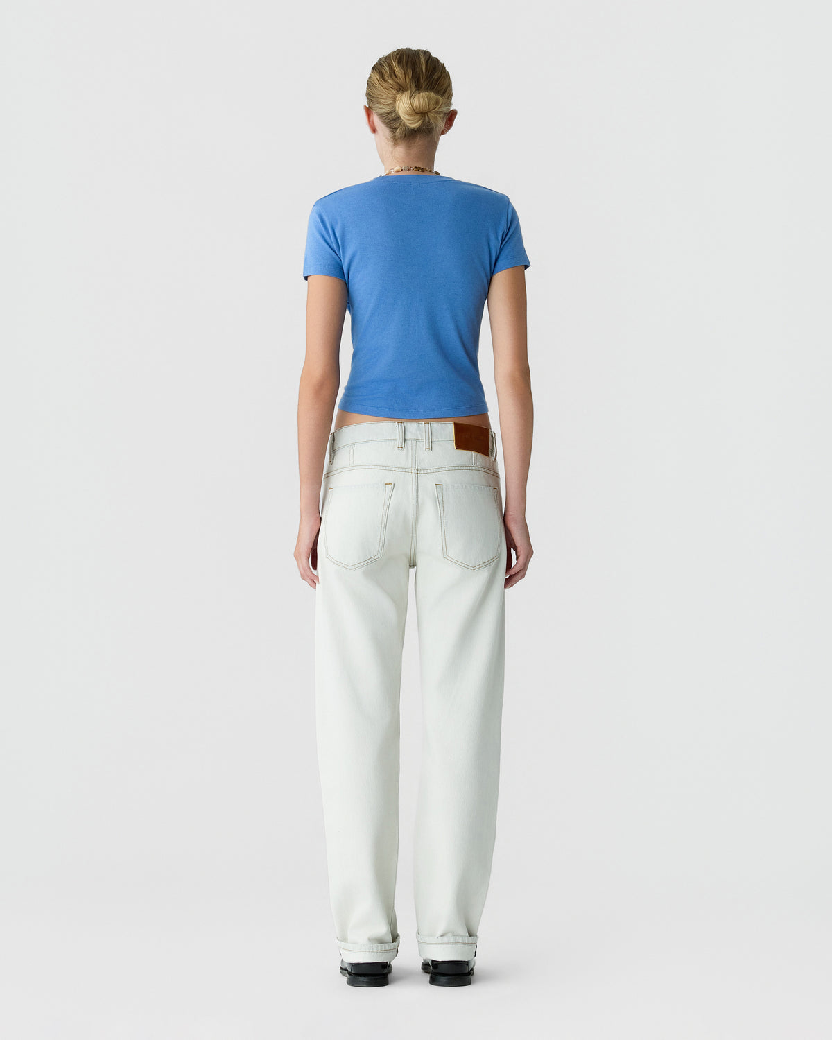 Back view of a person wearing a blue short sleeve T shirt, white wide leg jeans, and black shoes against a light gray background