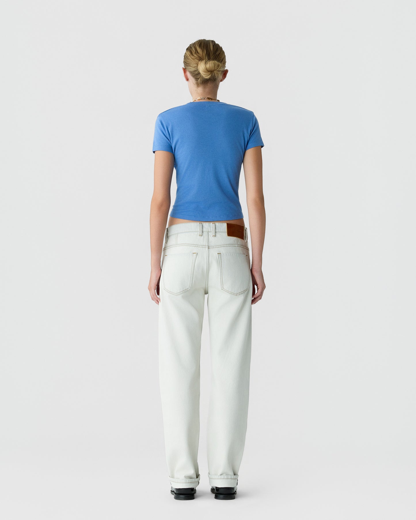 Back view of a model wearing a blue short sleeve shirt, white straight leg jeans, and black shoes against a light gray background