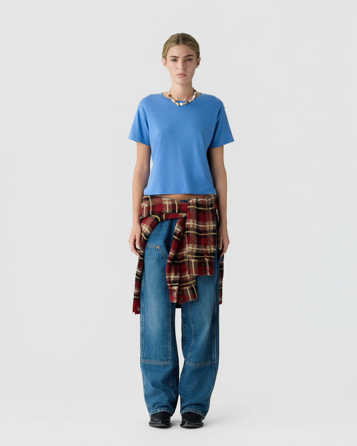 Model wearing a blue short sleeve shirt, relaxed blue jeans, black shoes, and a red plaid shirt tied around the waist against a light background