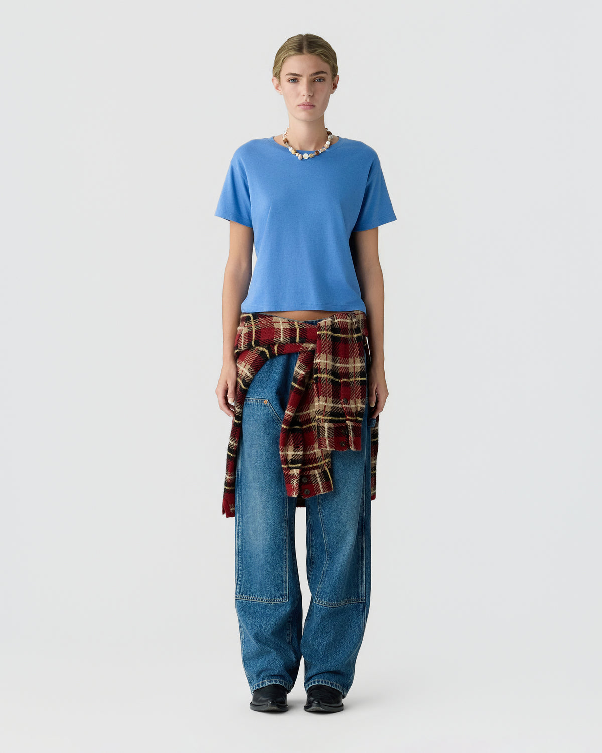Full-length studio portrait of a woman wearing a blue T-shirt, loose blue jeans, black shoes, and a red plaid shirt tied around her waist.