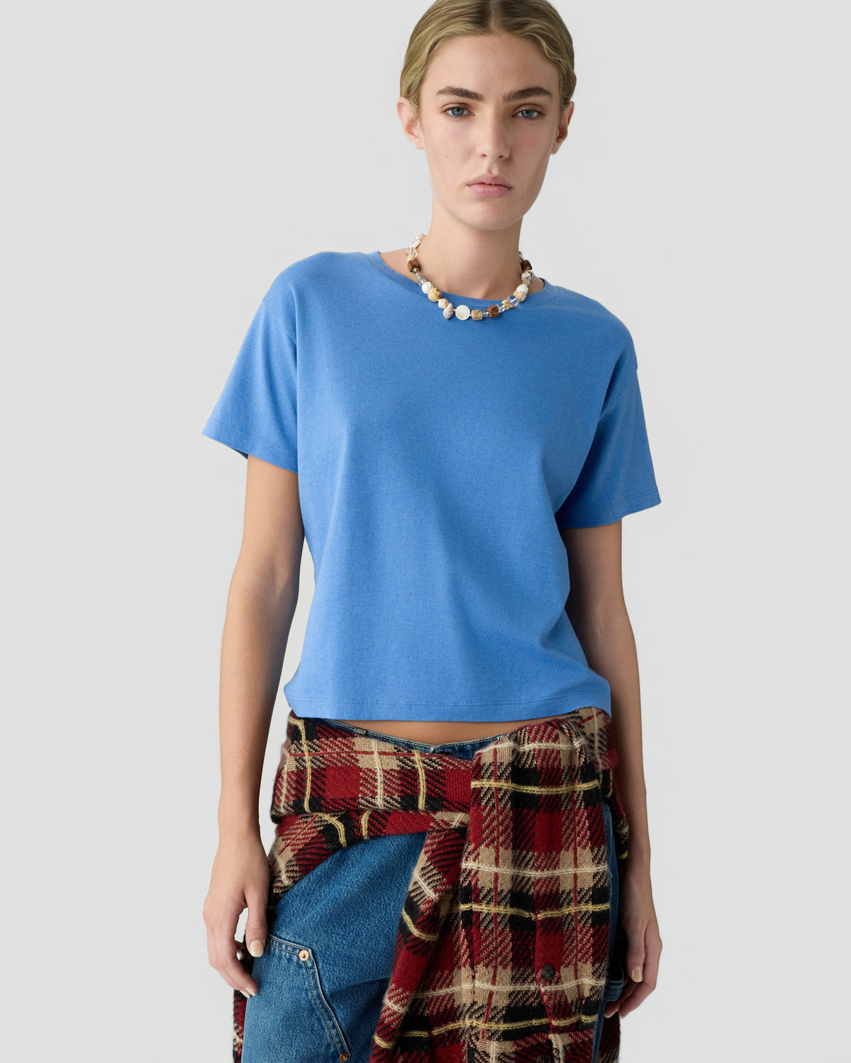 Model wearing a blue short sleeve cashmere top with a beaded necklace and red plaid trousers against a light gray background