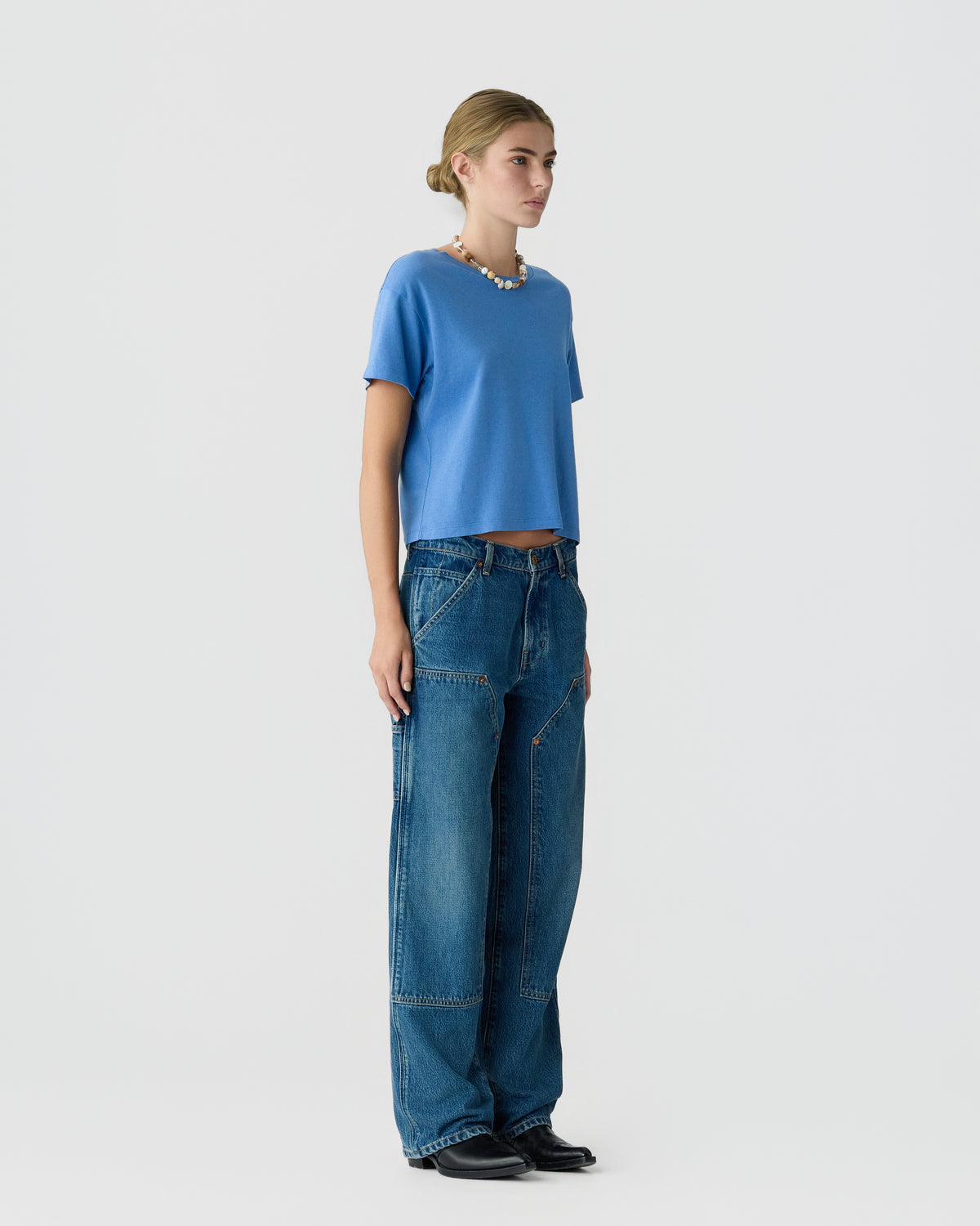 Woman wearing a blue short sleeve top with loose fit patchwork jeans and black shoes against a light gray studio background.