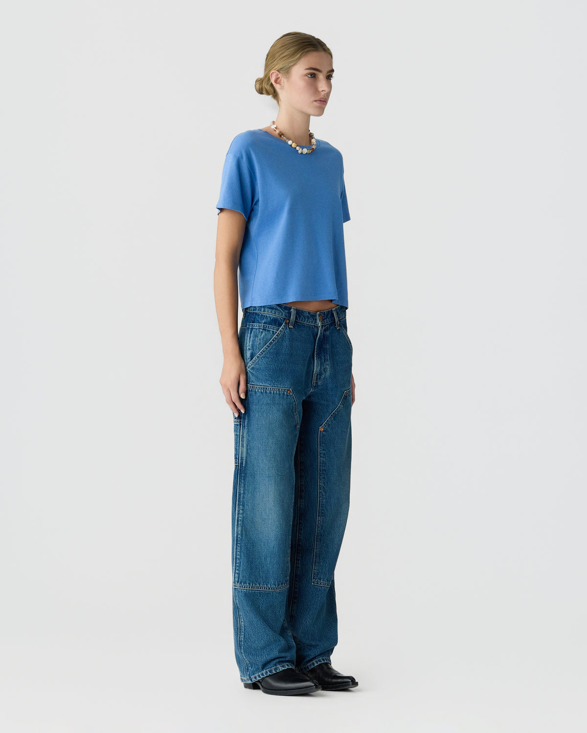 Woman standing against a light gray background wearing a blue cropped T-shirt wide-leg jeans a beaded necklace and black shoes