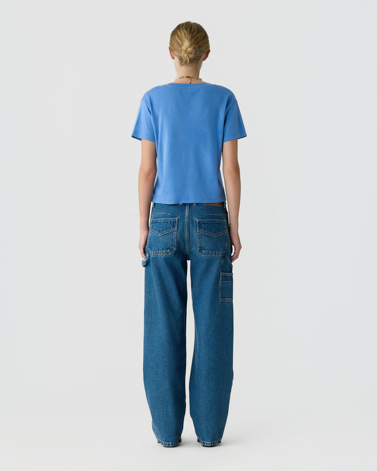 Rear view of a model wearing a blue short sleeve shirt and relaxed fit dark blue jeans with multiple utility pockets against a plain light background