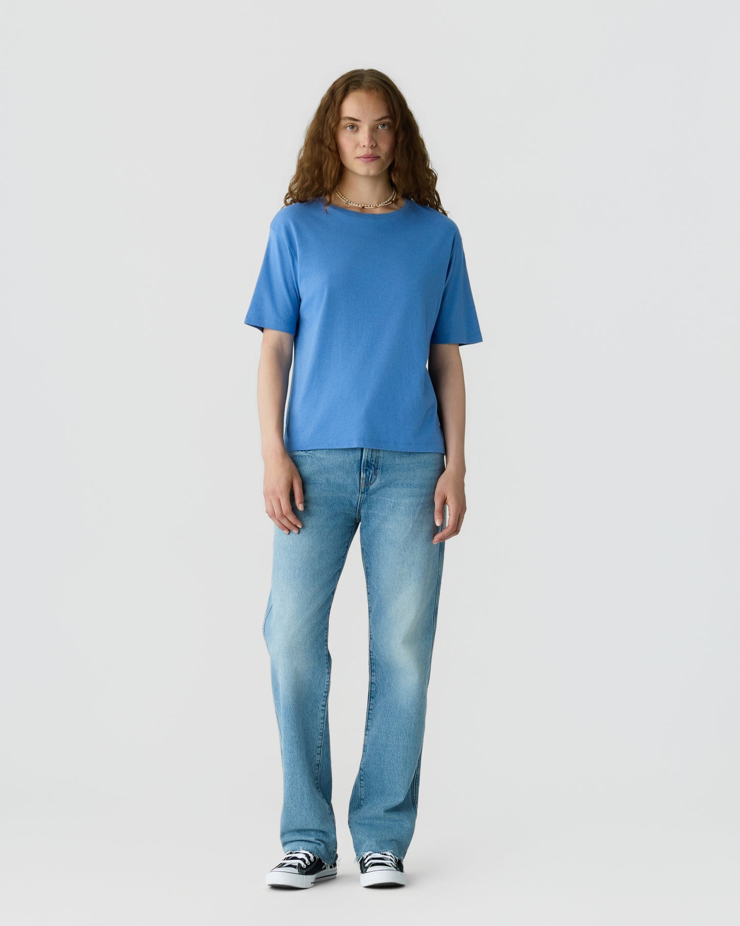 Woman with curly brown hair wearing a blue short sleeve shirt, light blue jeans, and black and white sneakers, standing against a light gray background