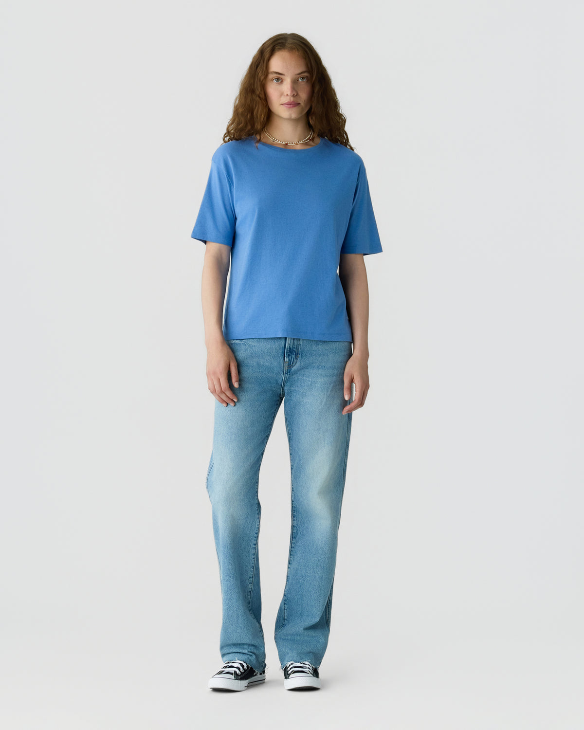 Woman with long brown hair wearing a blue short sleeve top with jeans and black and white sneakers standing against a light gray background