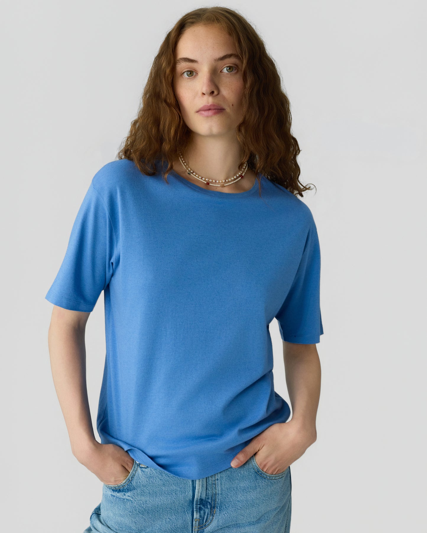 Woman with curly brown hair wearing a bright blue short sleeve top and light blue jeans while standing against a plain gray background