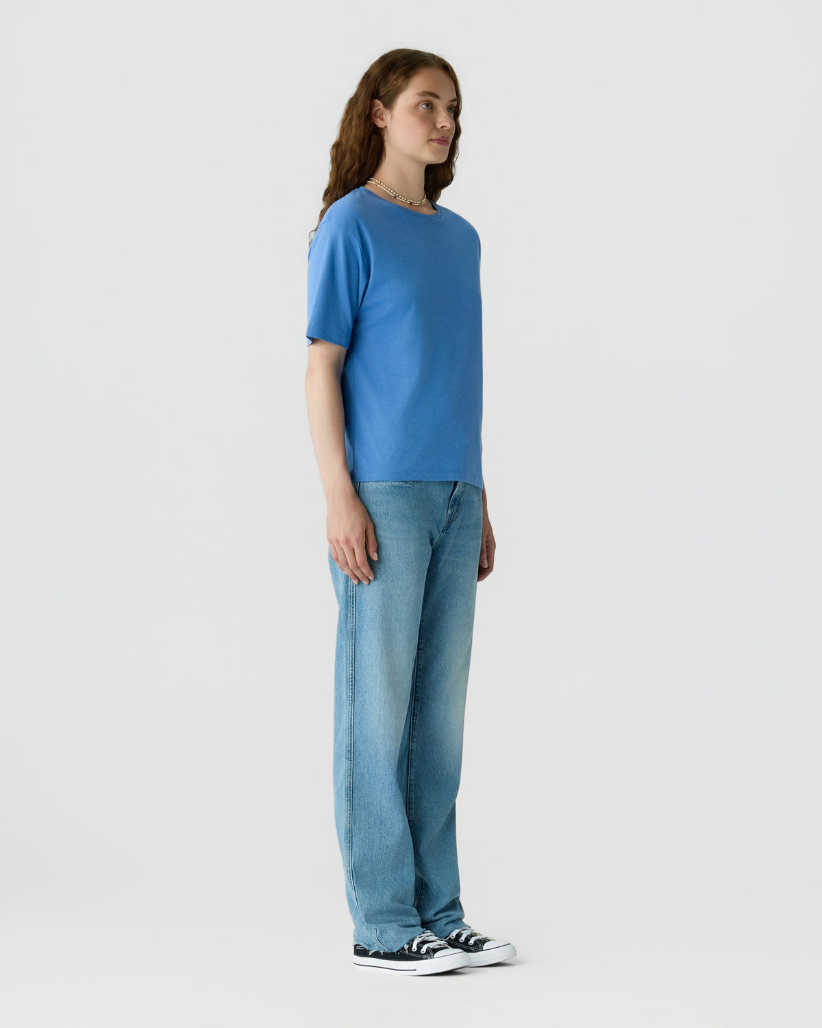 A woman stands in profile wearing a blue short sleeve top, light blue jeans, and black and white sneakers against a plain light background