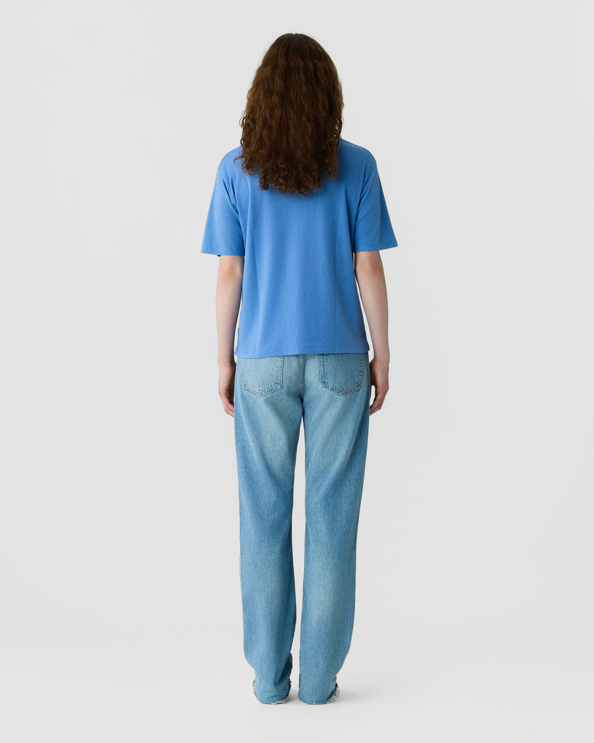 Back view of a person with long brown hair wearing a bright blue short sleeve shirt and relaxed light blue jeans against a plain light gray background
