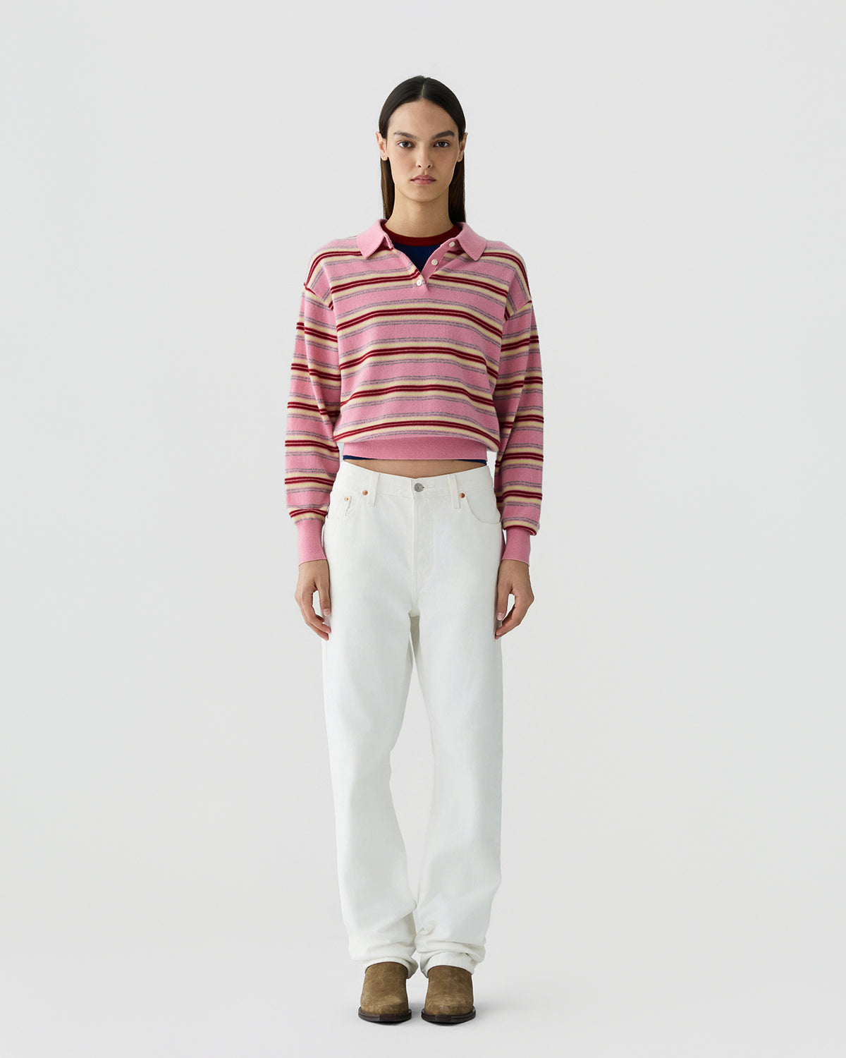 Woman standing facing forward wearing a pink striped knit top with a collar, paired with loose white pants and brown footwear, against a plain light background.