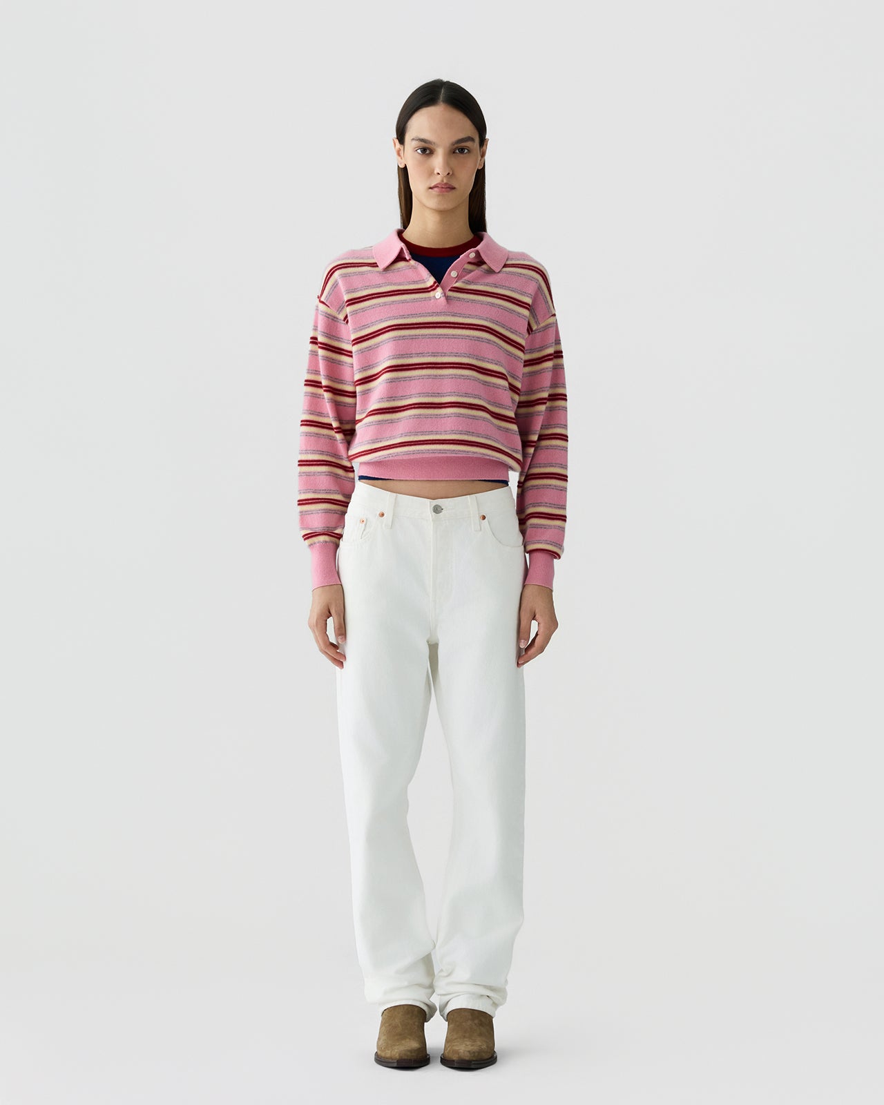 Woman standing facing forward wearing a pink striped knit top with a collar, paired with loose white pants and brown footwear, against a plain light background.