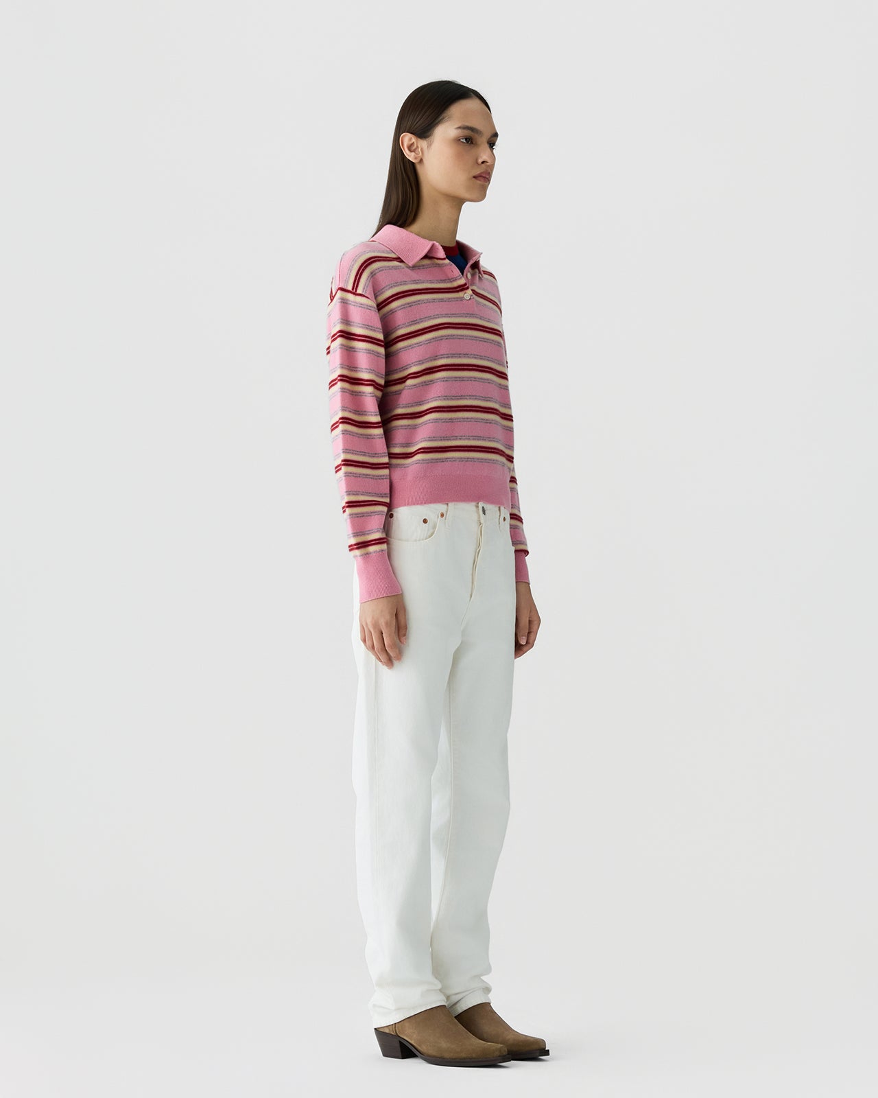 Woman standing in profile wearing a long-sleeve collared top with horizontal stripes in shades of pink and white high-waisted jeans, paired with tan heeled boots against a plain background.