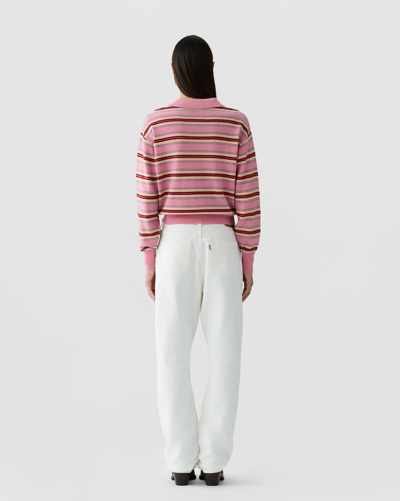 Woman seen from behind wearing a pink striped long-sleeve knit top with a collar, paired with high-waisted white pants and dark shoes, standing against a plain background.