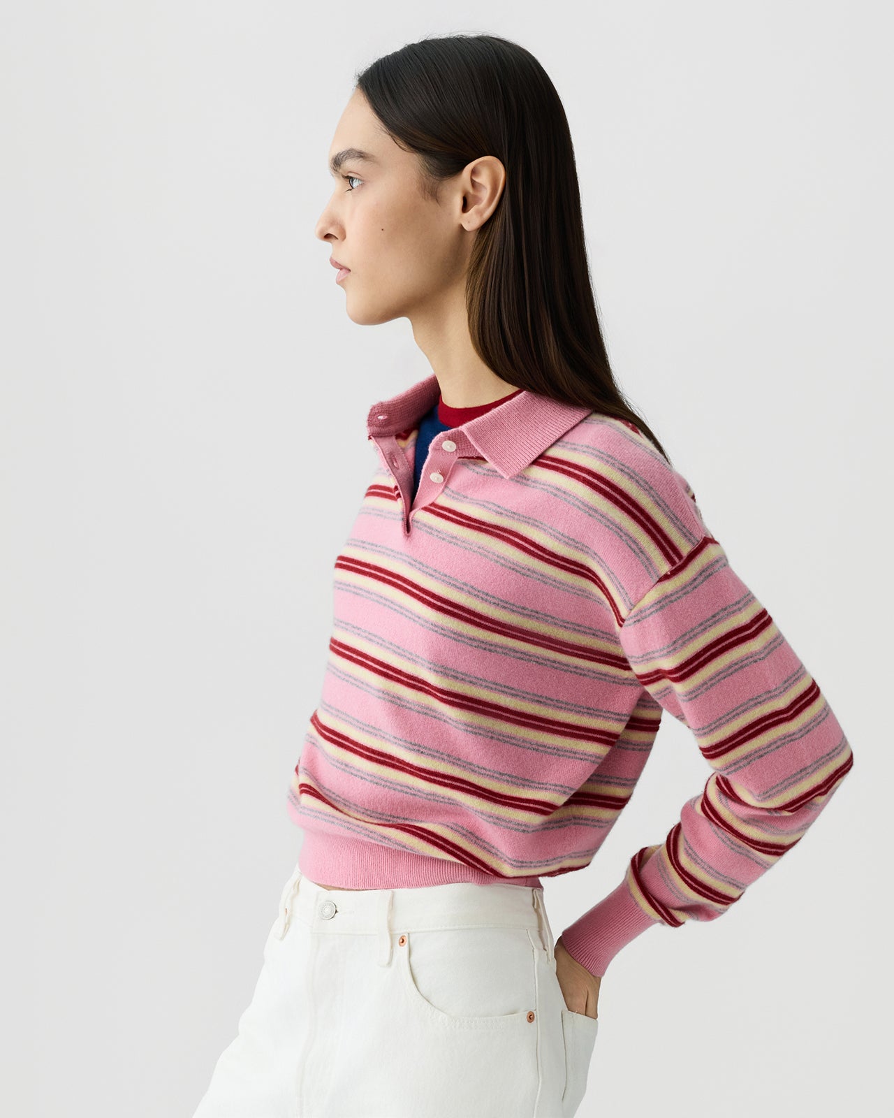 Woman shown in profile wearing a pink collared knit top with horizontal stripes in shades of red, cream, and light purple, paired with high-waisted white pants against a plain background.