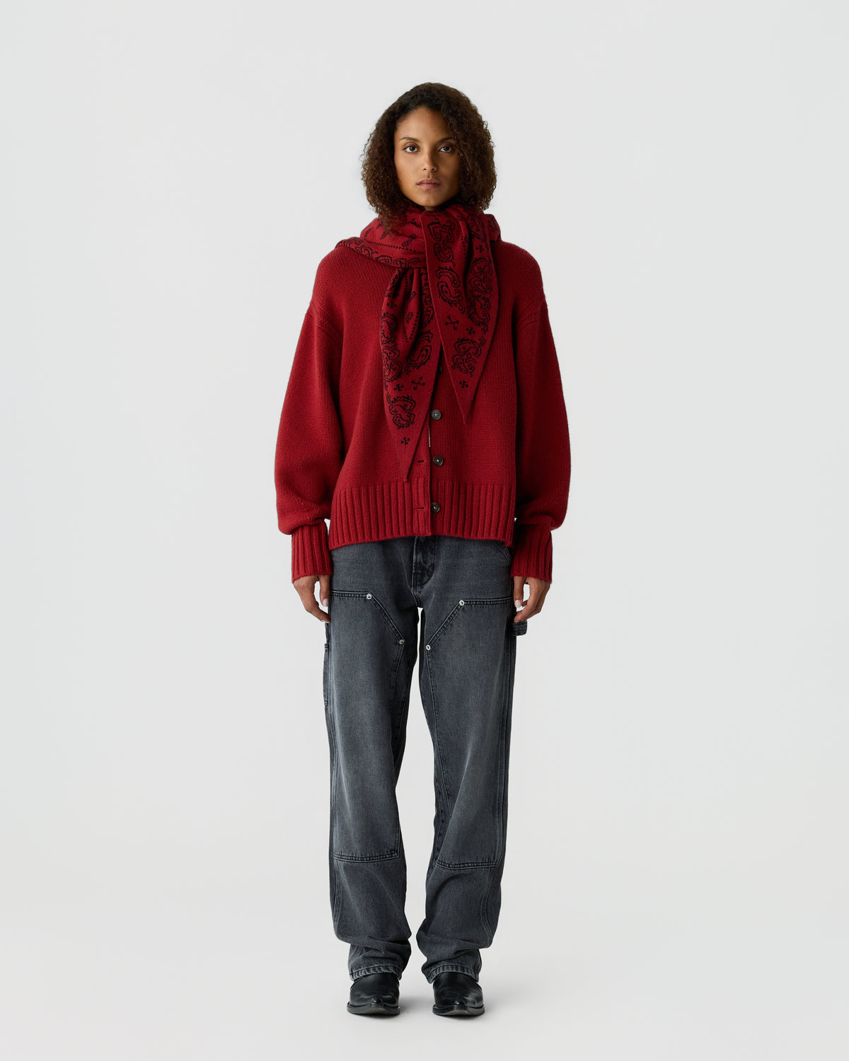 Woman standing against plain background wearing a red cardigan with large buttons, a matching red patterned scarf, faded black jeans, and black shoes.