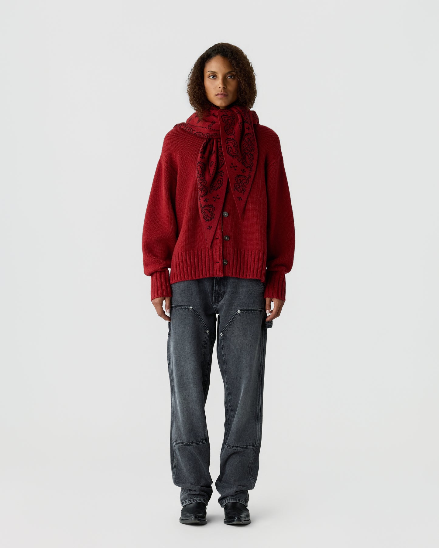 Woman standing against plain background wearing a red cardigan with large buttons, a matching red patterned scarf, faded black jeans, and black shoes.