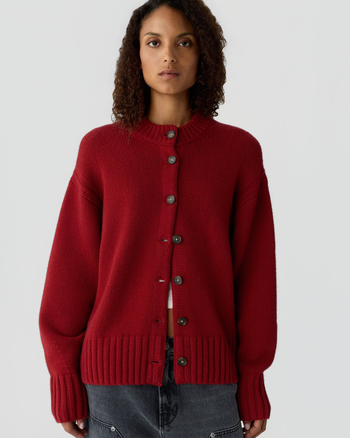Woman with curly hair wearing a deep red button-up cardigan sweater over a white top and dark gray pants against a plain light background.