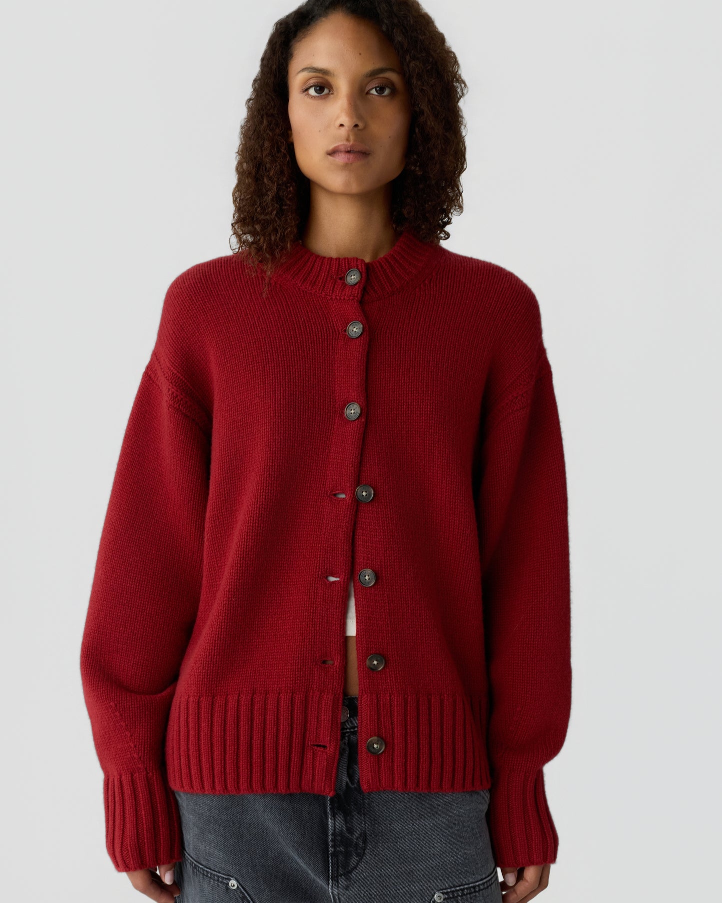 Woman with curly hair wearing a deep red button-up cardigan sweater over a white top and dark gray pants against a plain light background.