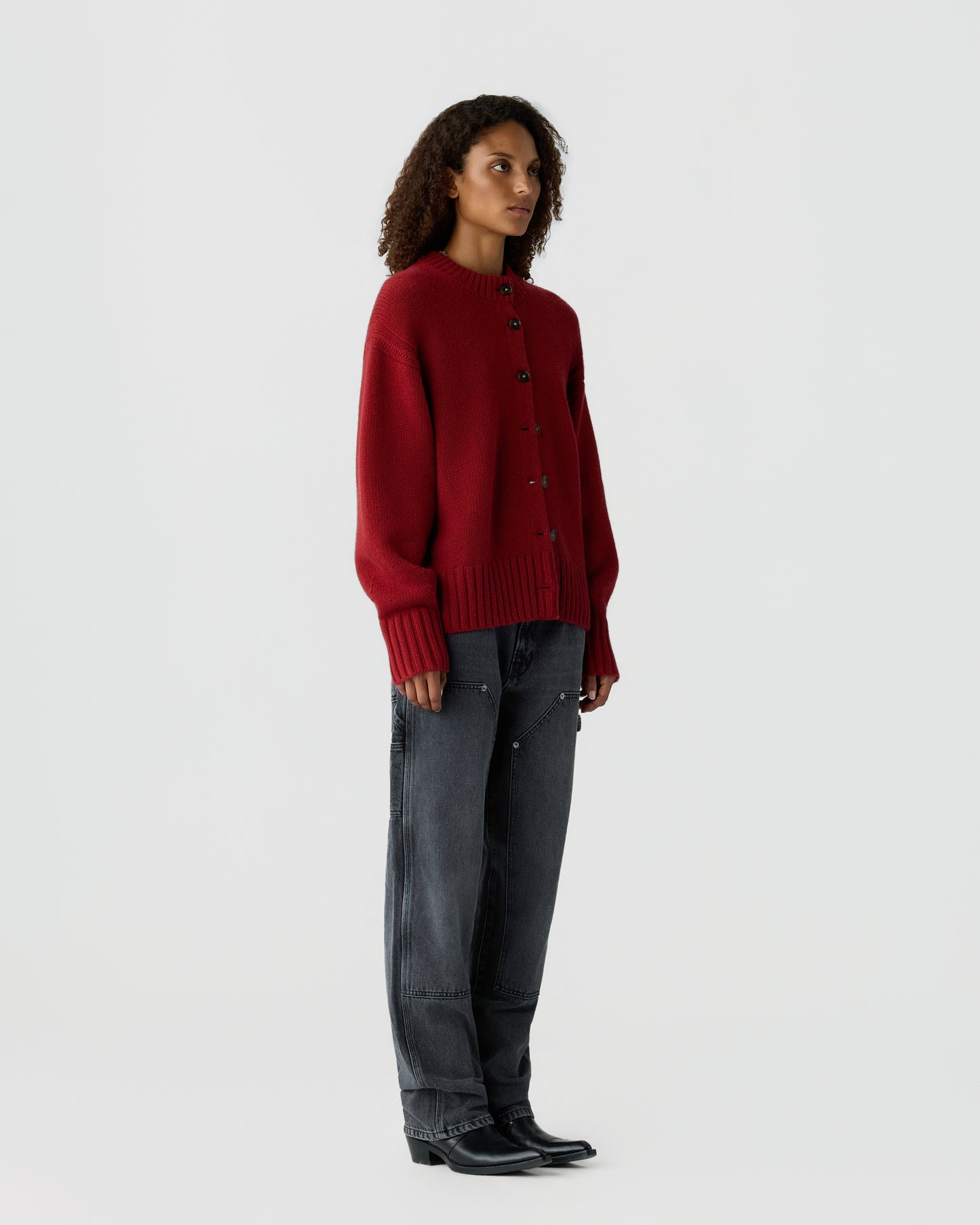Woman wearing a loose-fitting red buttoned sweater with long sleeves paired with dark gray wide-leg jeans and black boots, standing against a plain white background.