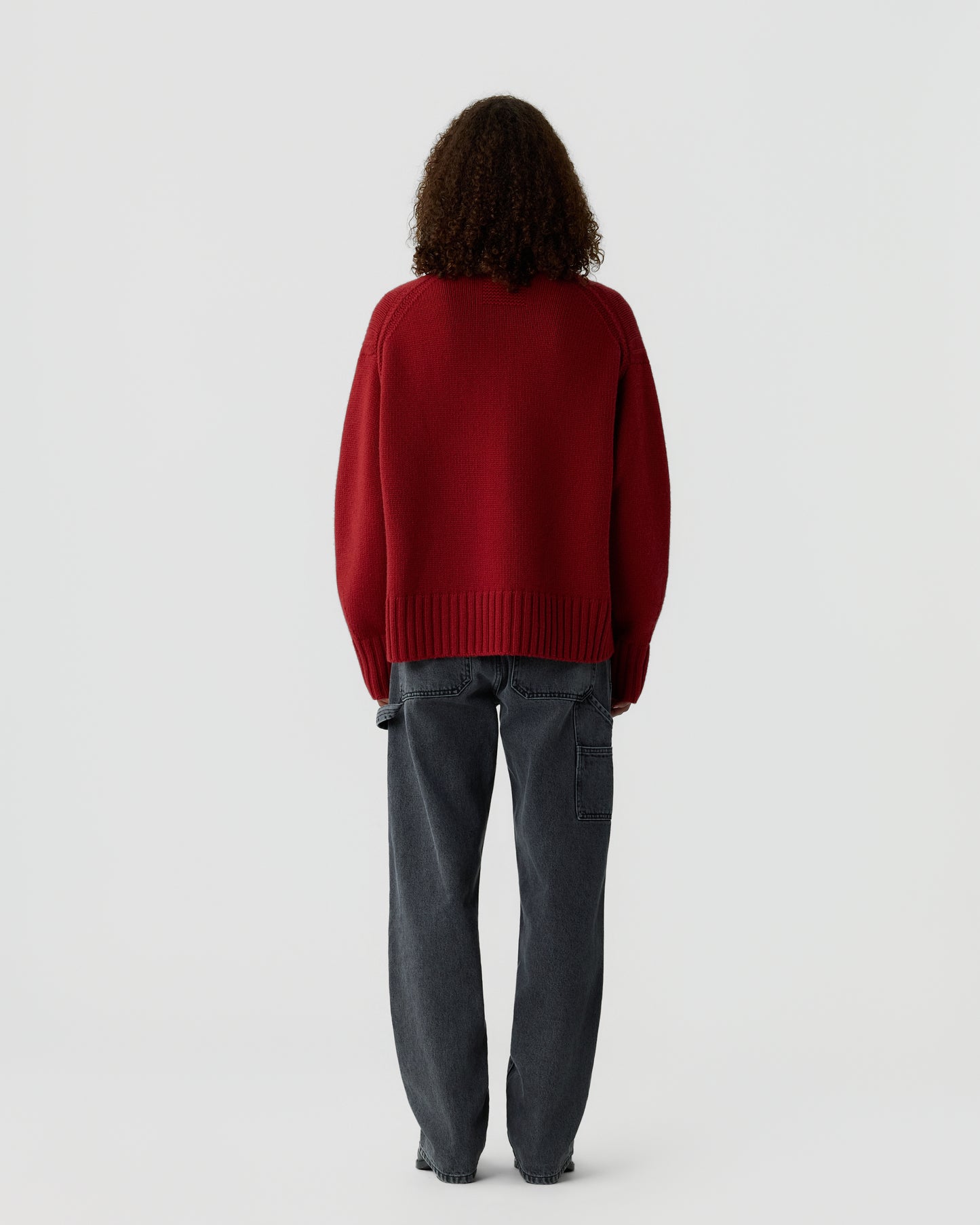 Person with curly hair wearing a loose red sweater and dark gray cargo pants, standing facing away against a plain light background.