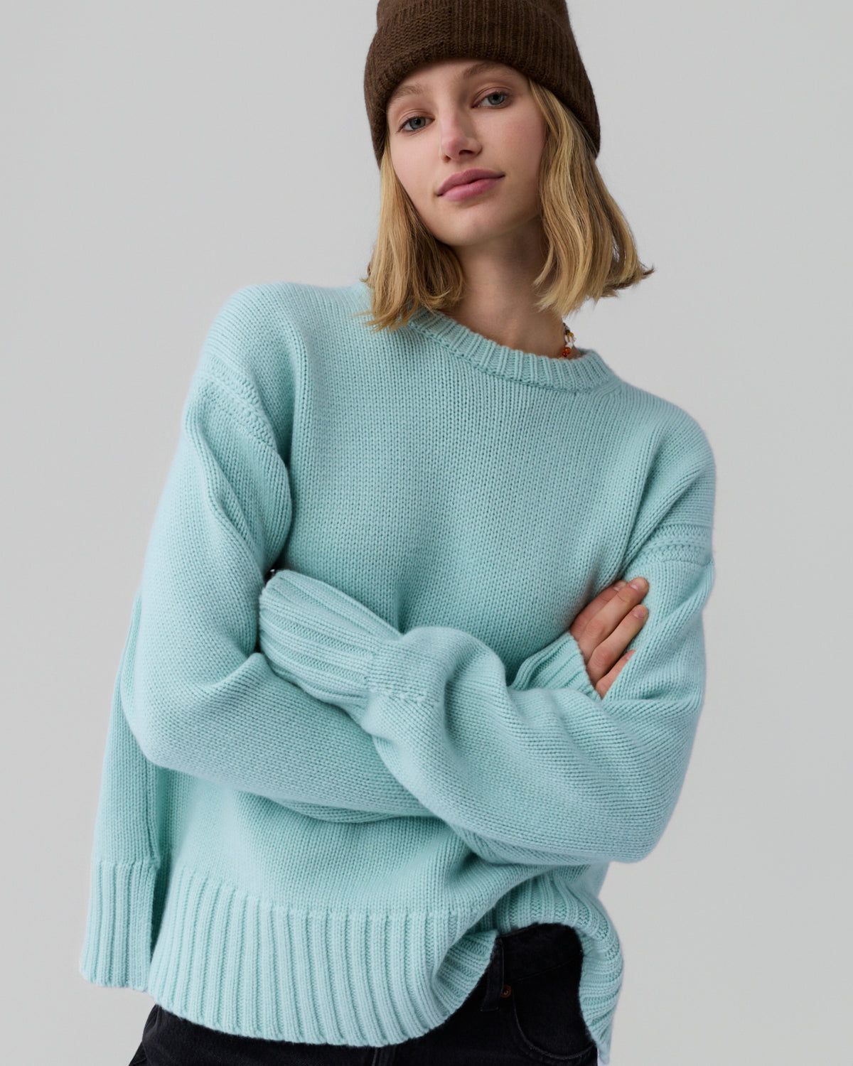 Young woman wearing a loose seafoam green knit sweater with wide ribbed cuffs and hem paired with black pants and a brown knit beanie against a plain light gray background.