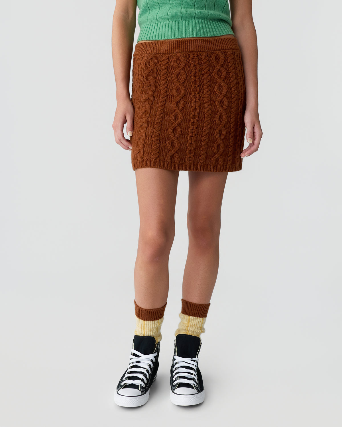 Lower half of a person wearing a brown cable-knit skirt, green ribbed top, yellow and brown socks, and black and white sneakers against a plain light background.