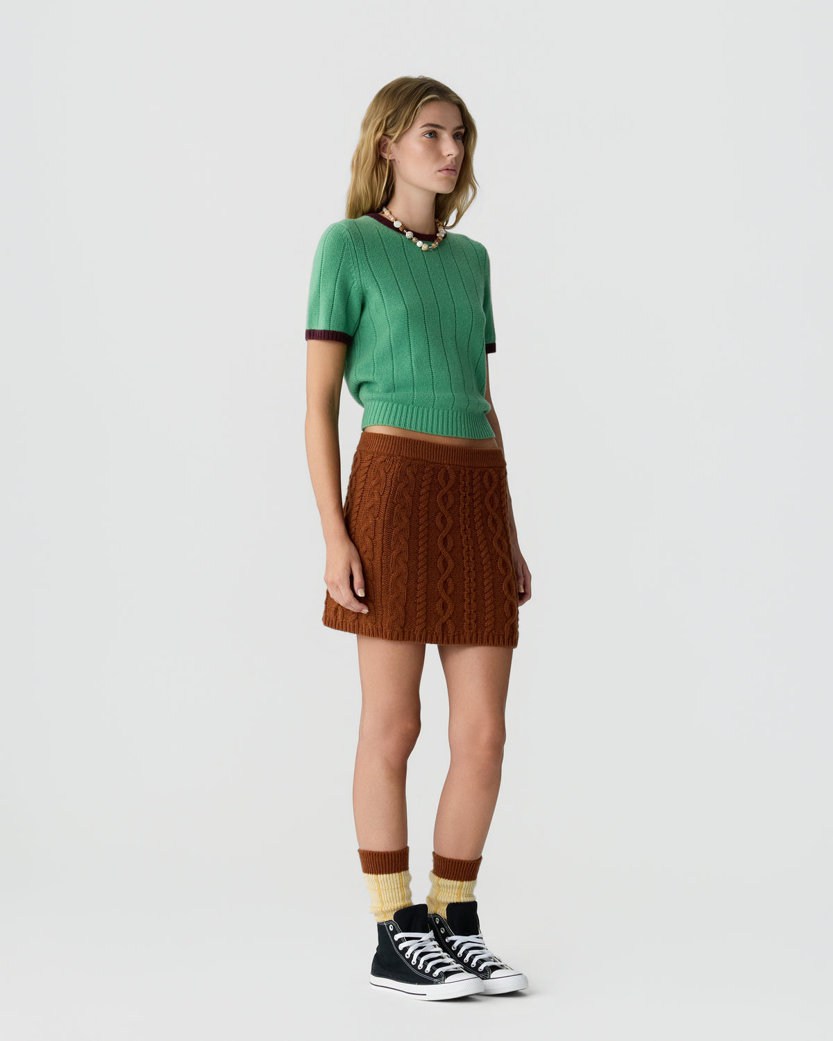 Woman wearing a green short-sleeve knit top with brown trim, a brown cable knit mini skirt, two-tone brown and cream socks, and black high-top sneakers, standing against a plain white background.