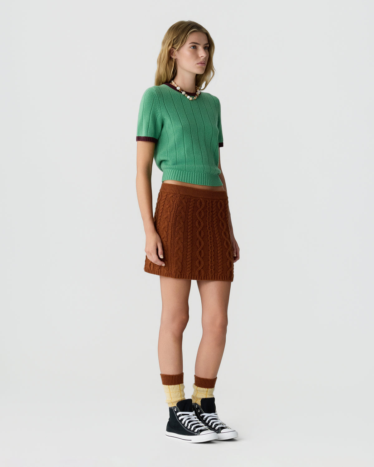 Model wearing a green ribbed knit sweater with burgundy trim, a brown cable knit skirt, striped socks, and black high top sneakers against a light background