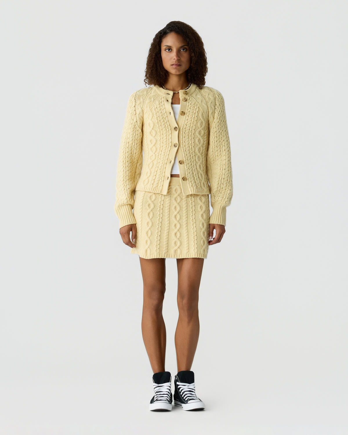 Woman wearing a cream cable knit cardigan with button front and matching skirt, styled with black and white sneakers against a light gray background