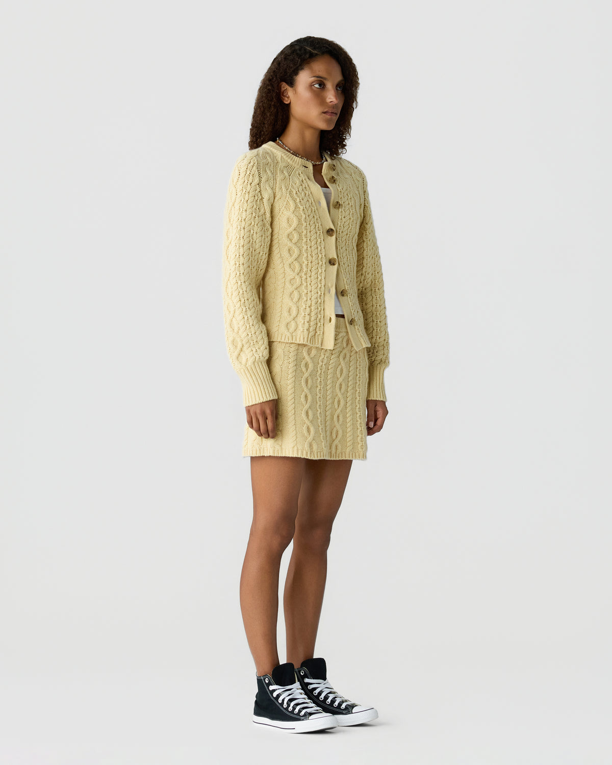 Model wearing a cream cable knit cardigan and matching textured mini skirt with black and white high top sneakers against a light gray background