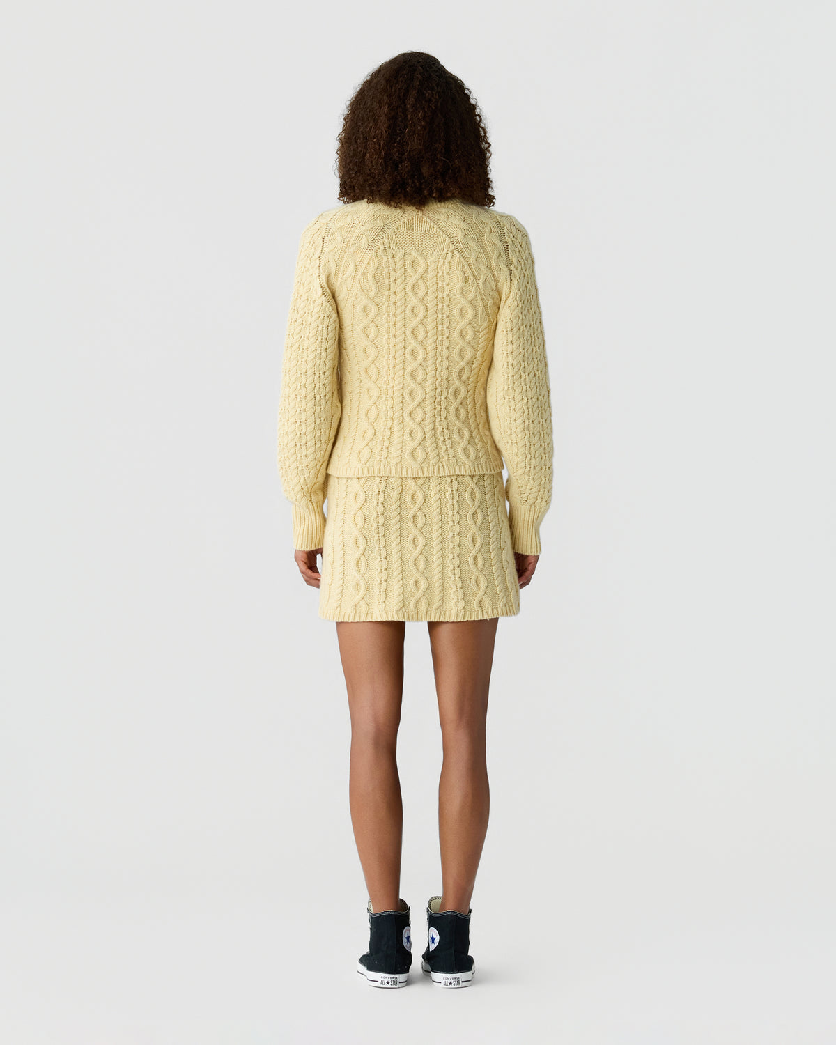 Back view of a person wearing a cream cable knit sweater with a matching skirt and black ankle boots against a light gray background