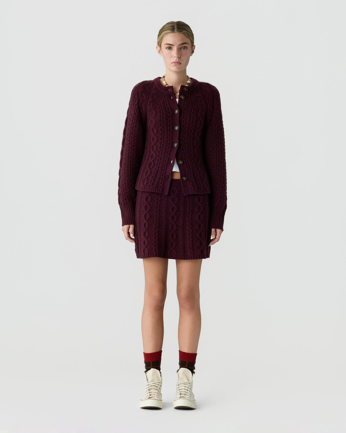 Model wearing a burgundy cable knit cardigan and matching mini skirt with ankle socks and cream sneakers against a light gray studio background