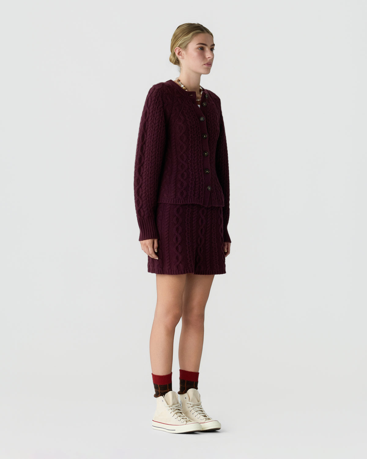 Model wearing a burgundy cable knit cardigan and matching skirt with red socks and white sneakers against a light gray background