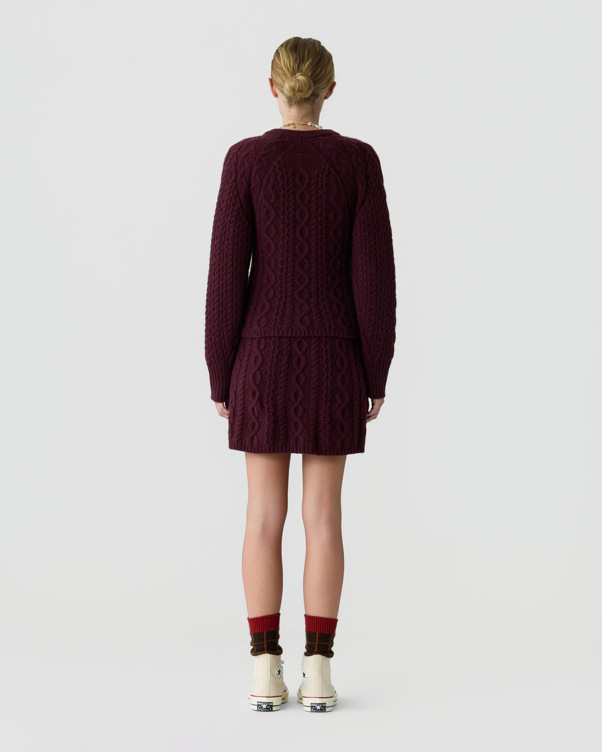 Back view of a model wearing a burgundy cable knit cardigan and matching skirt with red socks and cream shoes against a light gray background