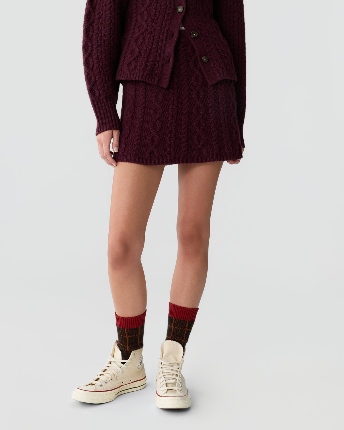 Lower half of a person wearing a burgundy cable-knit skirt and matching cardigan with patterned dark red and black socks and white high-top sneakers against a plain light background.