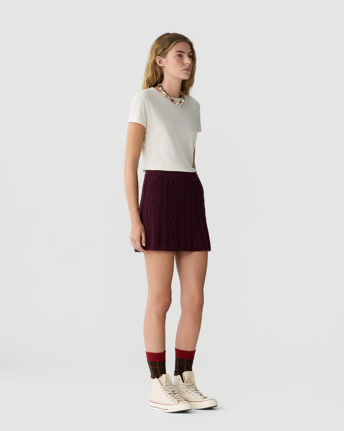 Young woman standing against a plain background wearing a white short-sleeve top, a dark purple patterned mini skirt, red and black checkered socks, and off-white high-top sneakers.