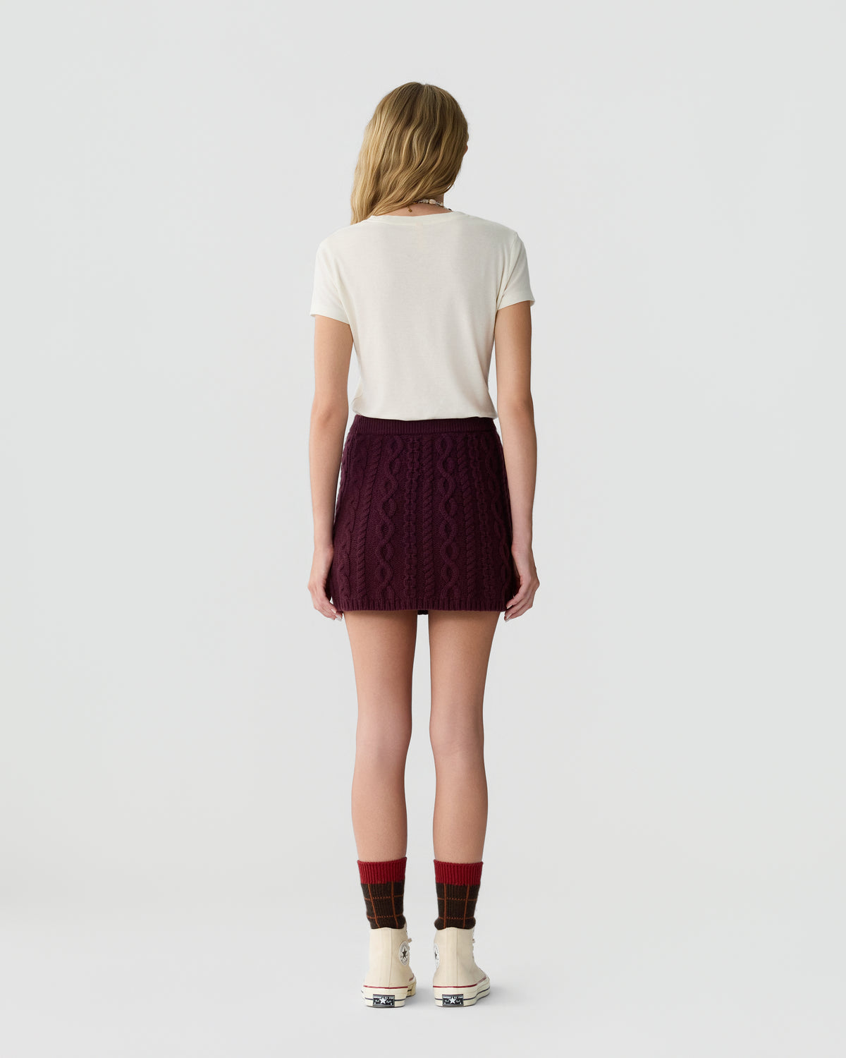 Woman wearing a white short-sleeve top and a burgundy cable knit mini skirt, paired with red and brown plaid socks and white high-top sneakers, standing against a plain background.