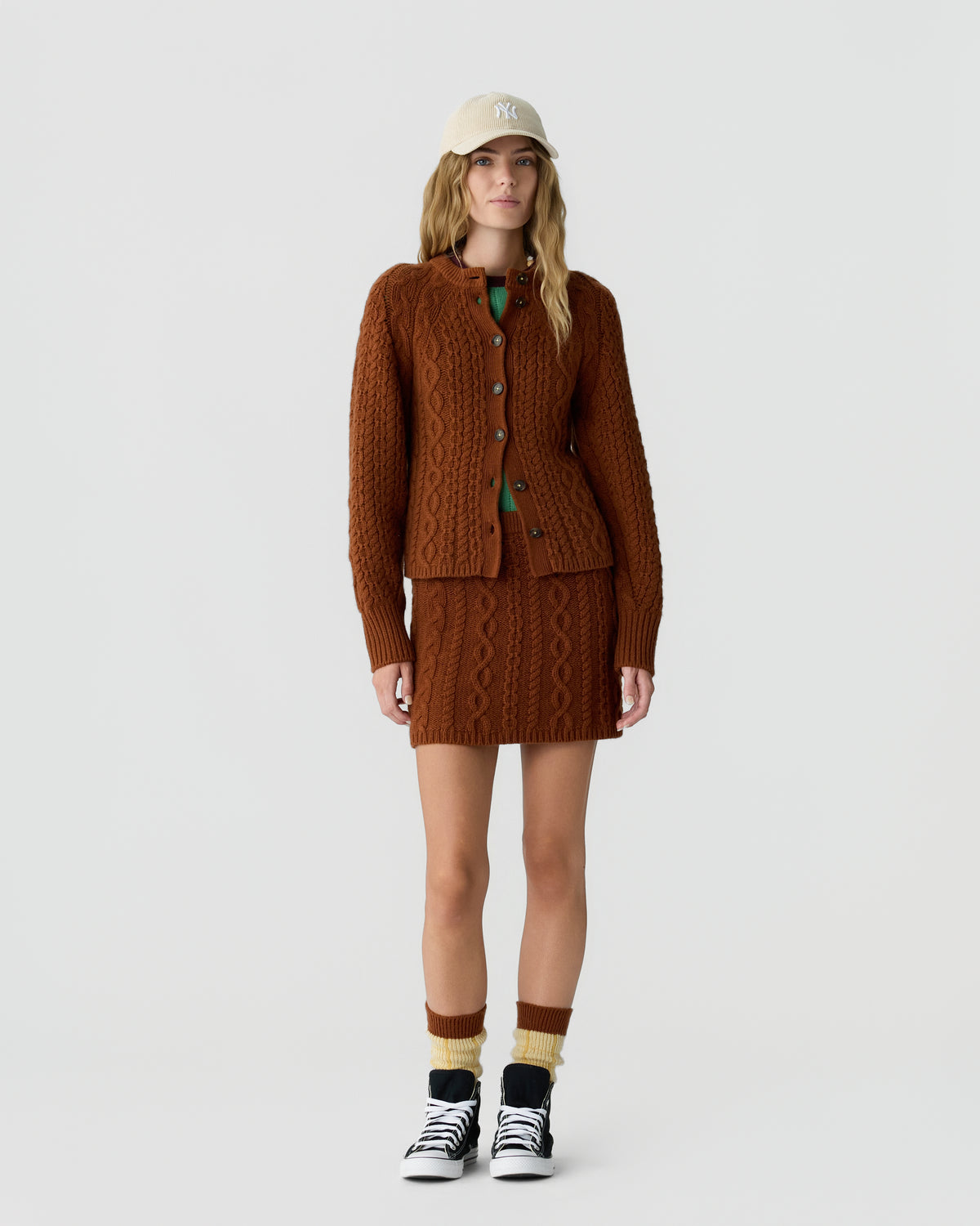 Woman wearing a brown cable knit cardigan and matching mini skirt outfit with multicolored socks, black and white sneakers, and a beige cap, standing against a plain background.