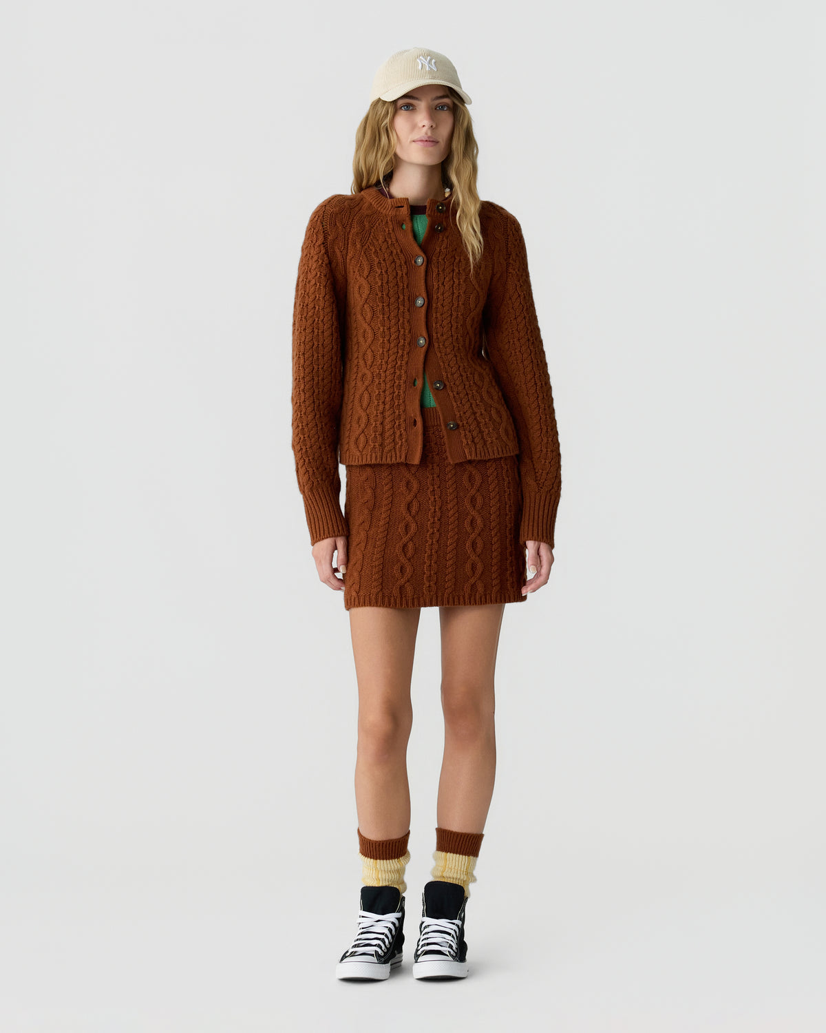 Model wearing a brown cable knit cardigan and matching skirt with a beige cap yellow socks and black and white sneakers on a light background