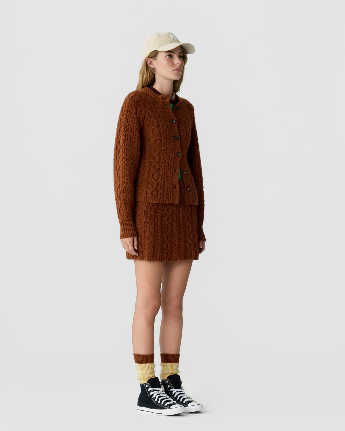 Woman wearing a brown cable-knit cardigan and matching skirt, cream and brown socks, black high-top sneakers, and a beige baseball cap against a plain light background.