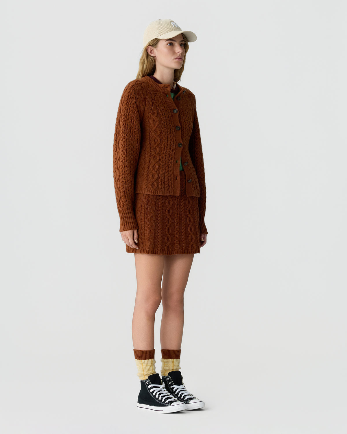 Woman wearing a brown cable knit cardigan and matching skirt with a beige cap striped socks and black high top sneakers against a light gray background